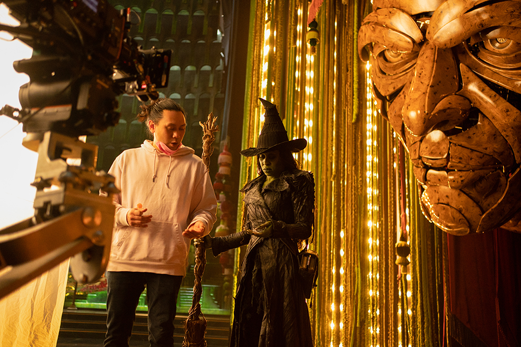 Director Jon M. Chu discusses a scene with Cynthia Erivo with the puppet of the Wizard of Oz’s head hanging in behind them.